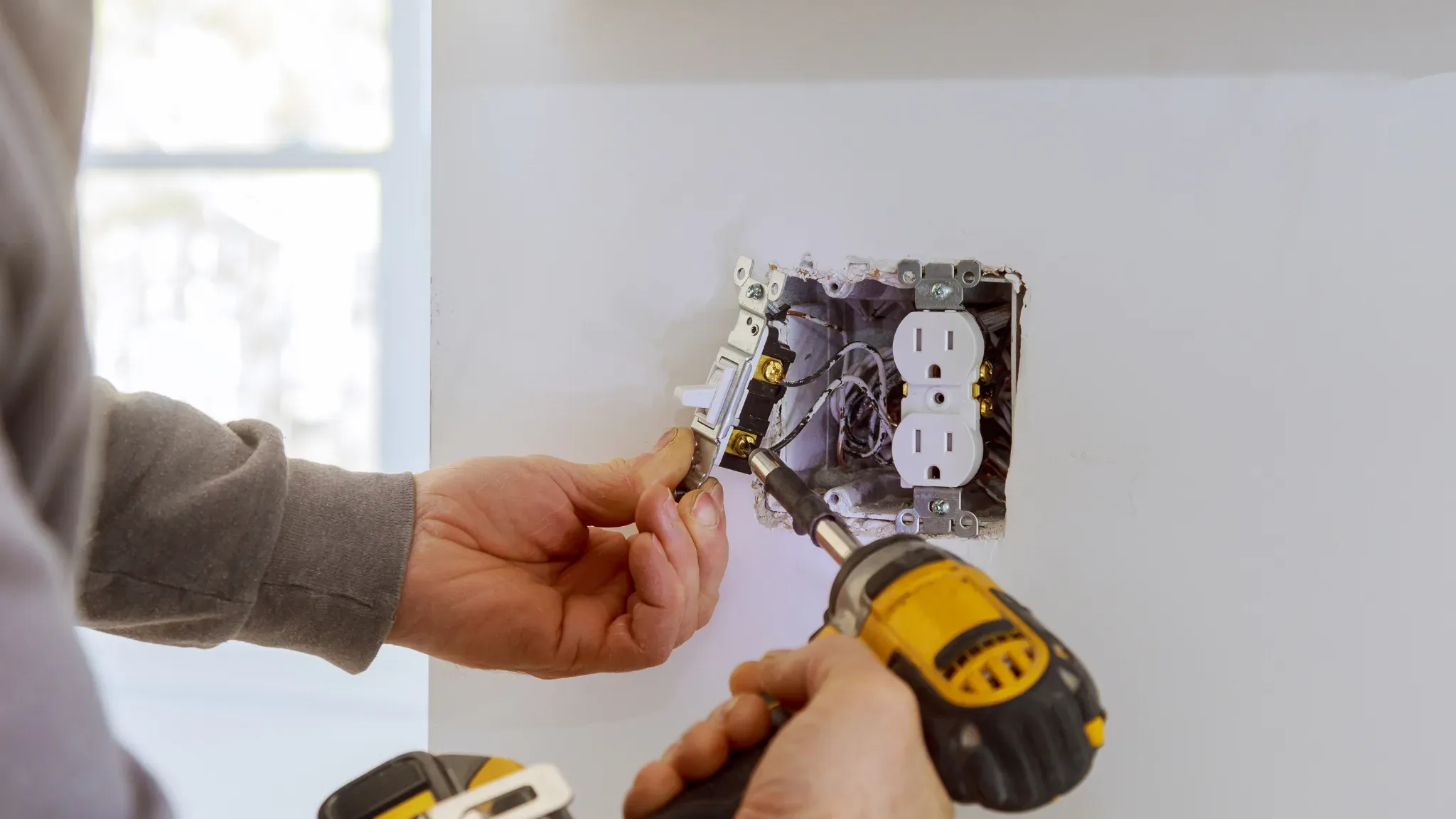 Close-up of an electrician's hands installing a white duplex electrical outlet into a wall box. One gray-sleeved hand holds t