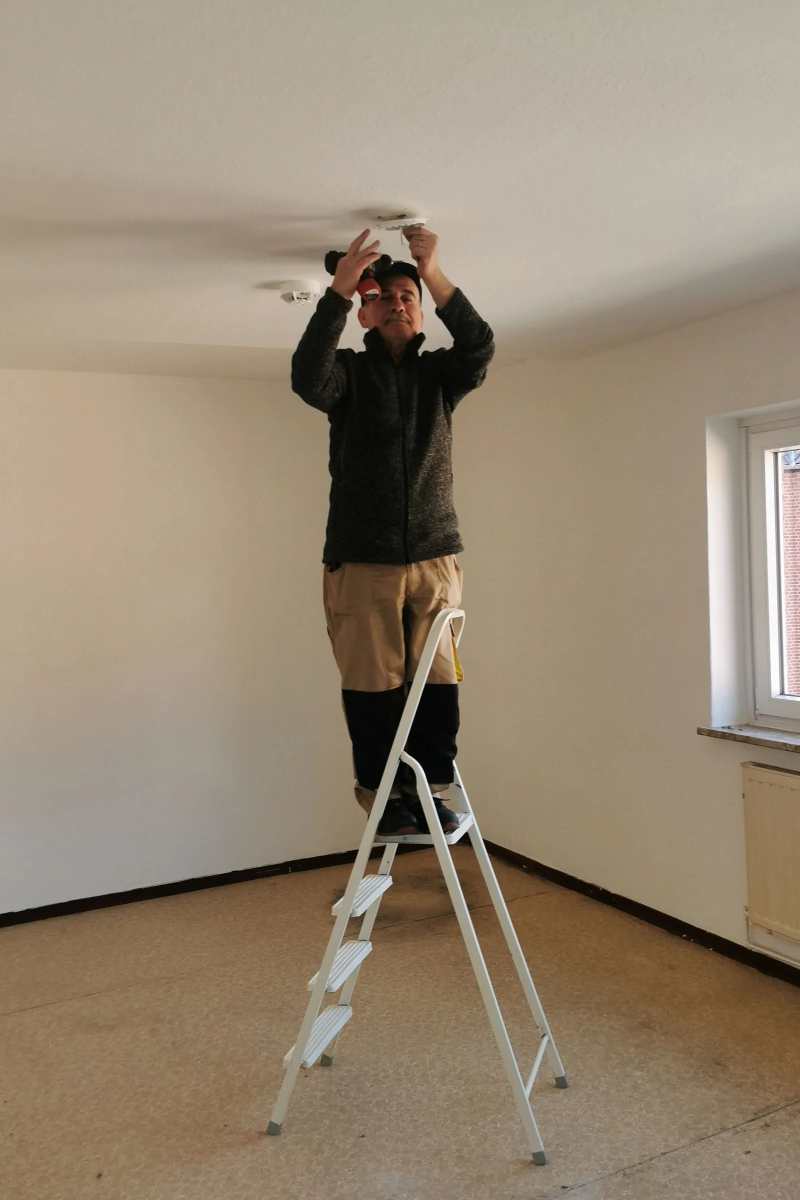 Electrician on ladder installing ceiling fixture in residential interior.